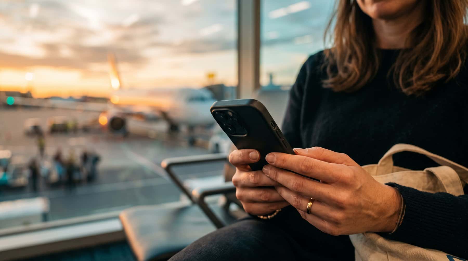 Hands holding a smartphone in an airport gate area with a plane outside at sunset